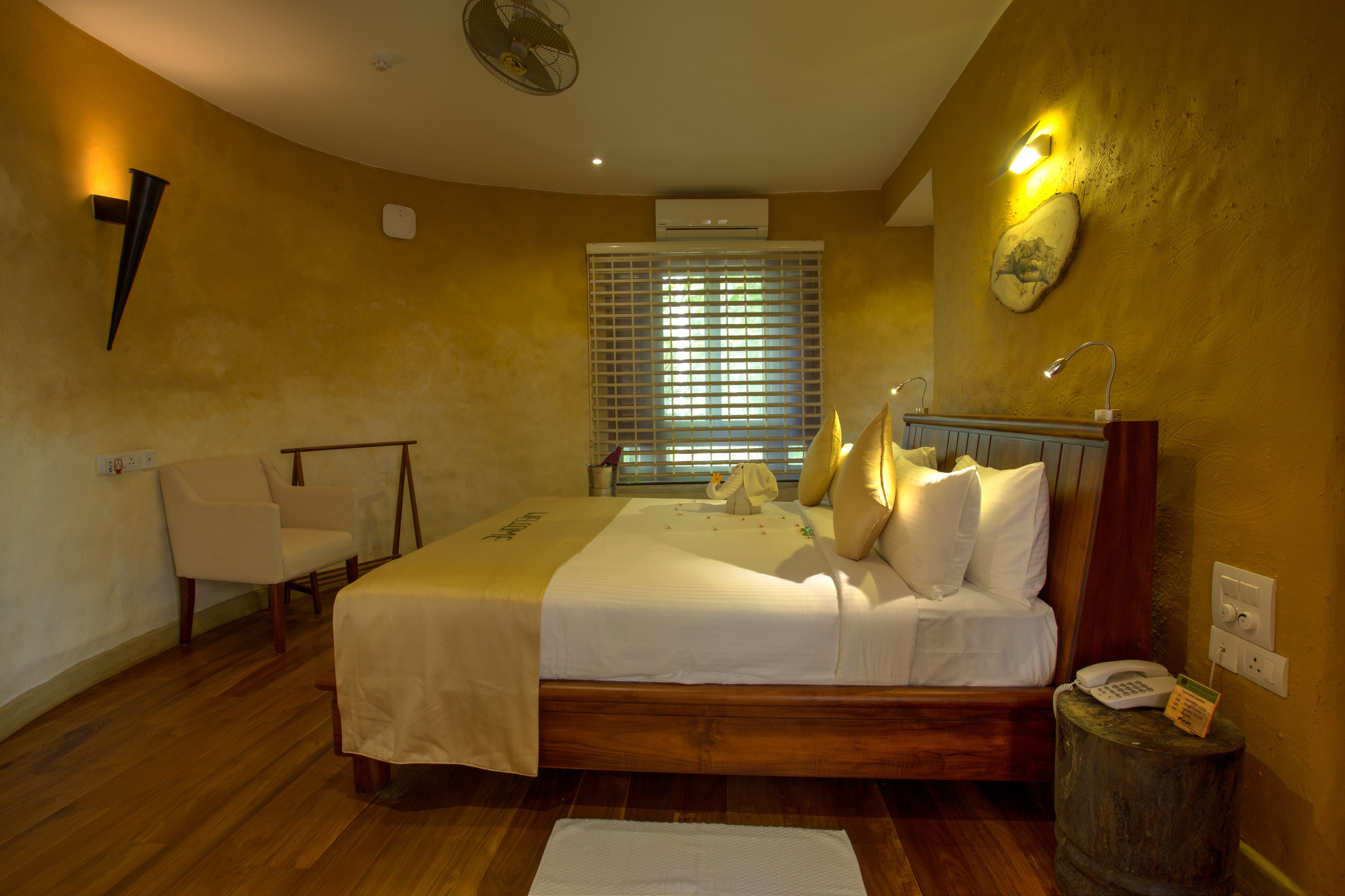 A bedroom with a bed, a chair, a bedside table, an air conditioner, a wall-mounted fan, and a window with blinds.