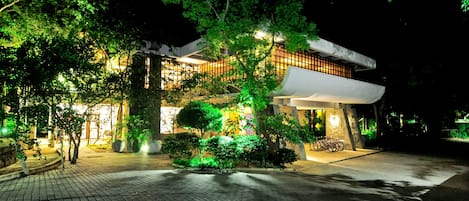 A modern building with a brick pathway leading to the entrance, surrounded by lush greenery and trees.