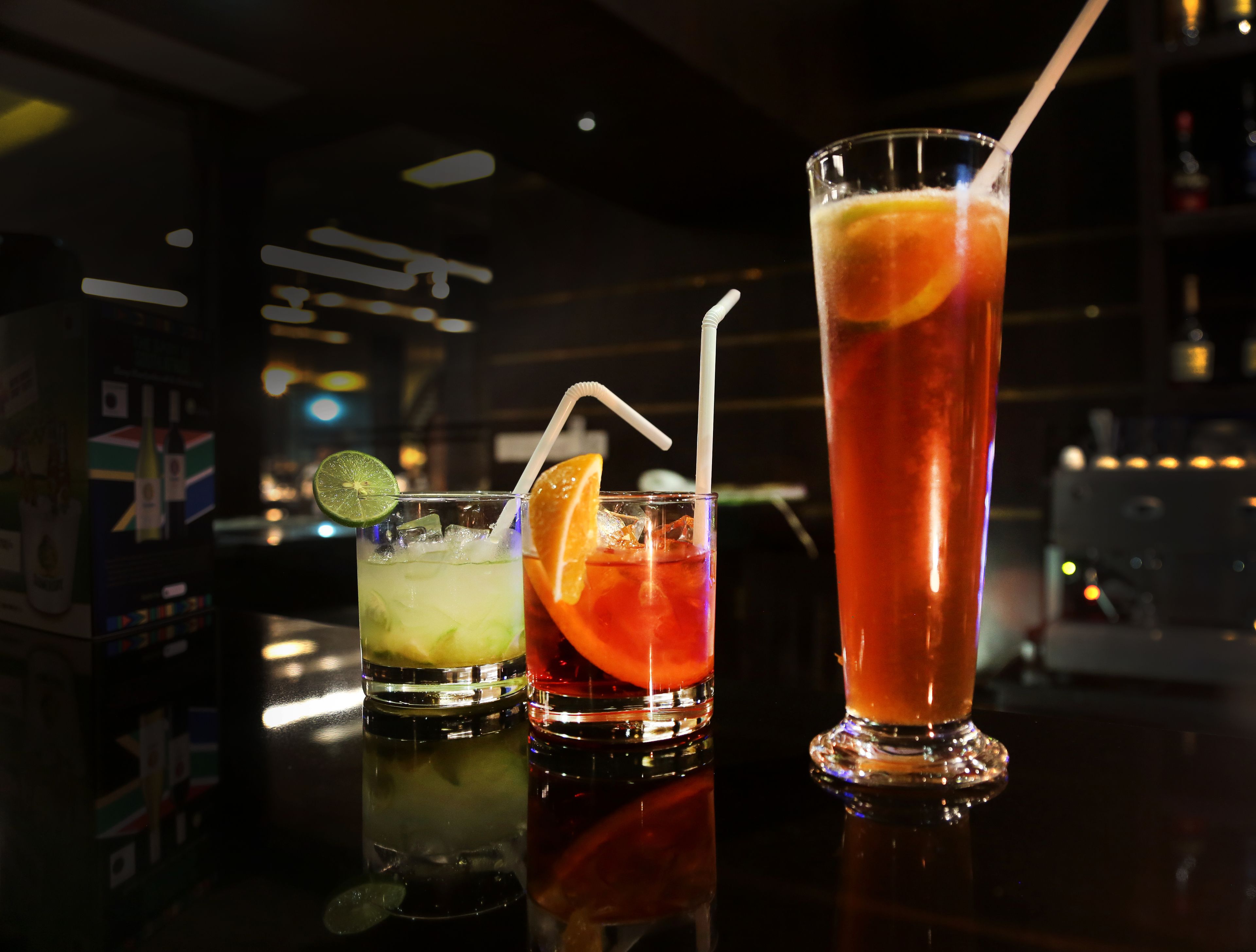 Three cocktails on a bar counter with a dark background and some illuminated signs.