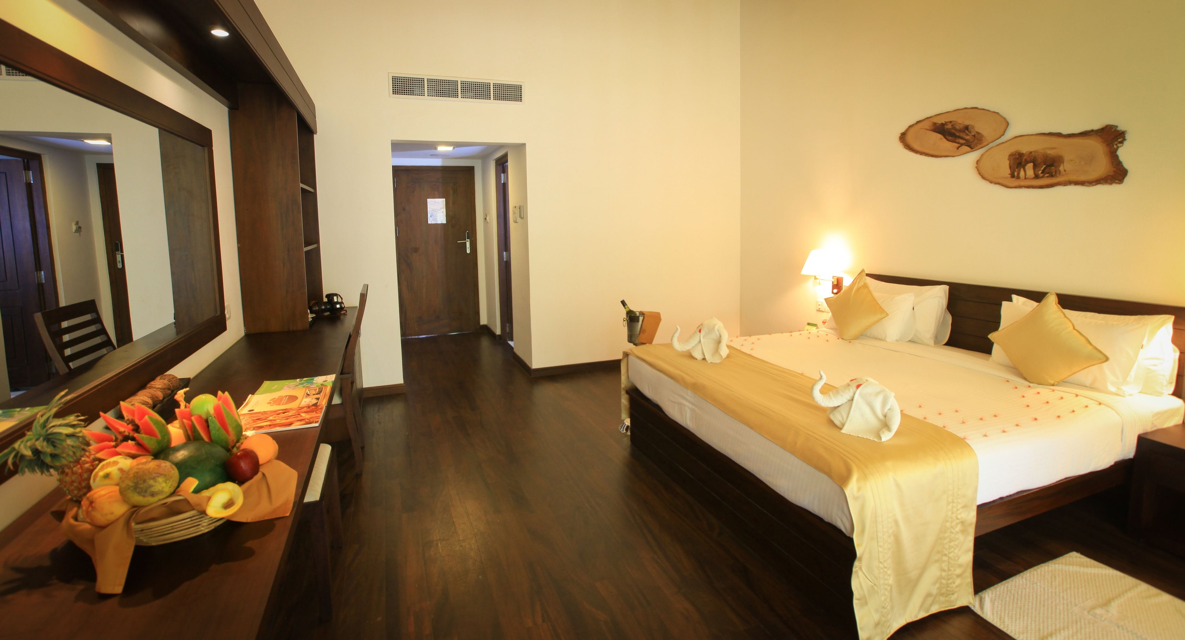 Habitación de hotel con piso de madera, cama con ropa blanca y un plaid dorado, una cesta de frutas y una lámpara fijada a la pared.