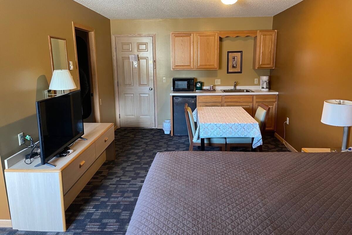 A hotel room with a bed, a TV, a desk, a chair, a small kitchenette, and a door.