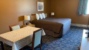 Habitación de hotel con una cama, una mesa con mantel floral, una silla, una lámpara de noche y un cuadro en la pared.