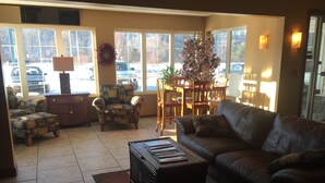 A living room with a leather sofa, patterned armchair, and a coffee table with magazines. There is a dining table with chairs and a potted plant. - Lakeside Motor Inn (Gladstone)