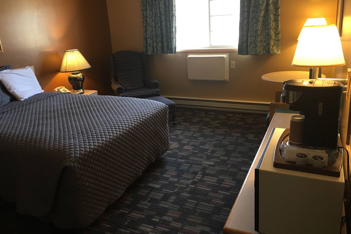 A hotel room with a bed, a chair, a desk with a coffee maker, and a window with curtains.