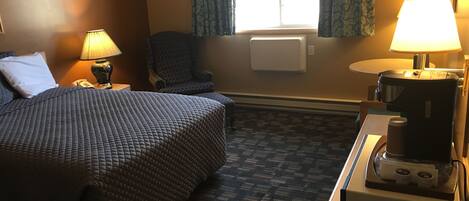 A hotel room with a bed, a chair, a desk with a coffee maker, and a window with curtains.
