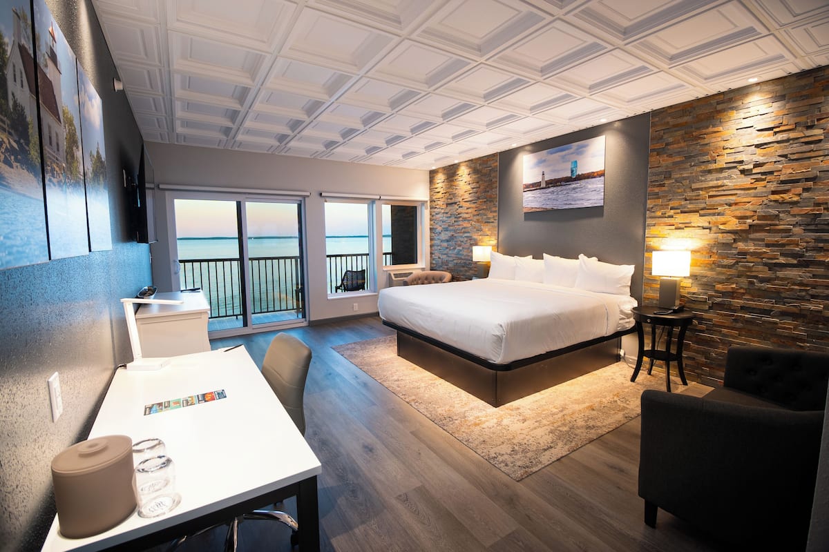 A modern hotel room with a large bed, a desk, a chair, a small table, and a view of the sea through the balcony doors.