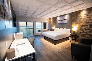 A modern hotel room with a large bed, a desk, a chair, a small table, and a view of the sea through the balcony doors.