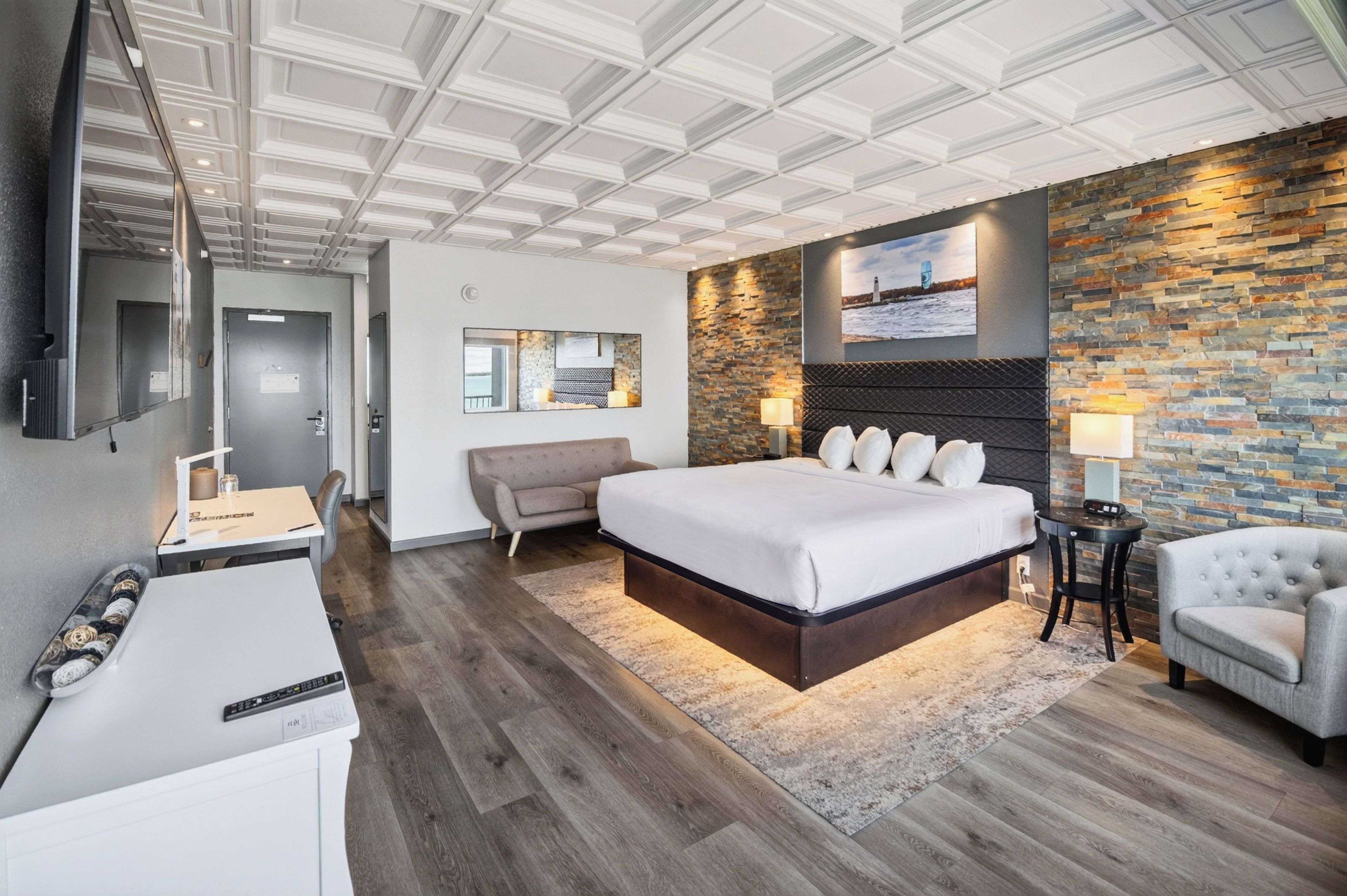 A modern hotel room with a large bed, a seating area, a TV, and a stone accent wall.