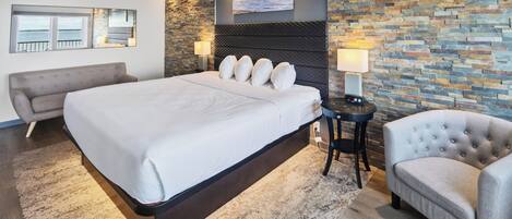 A modern hotel room with a large bed, a grey armchair, a small round table, and a stone accent wall.