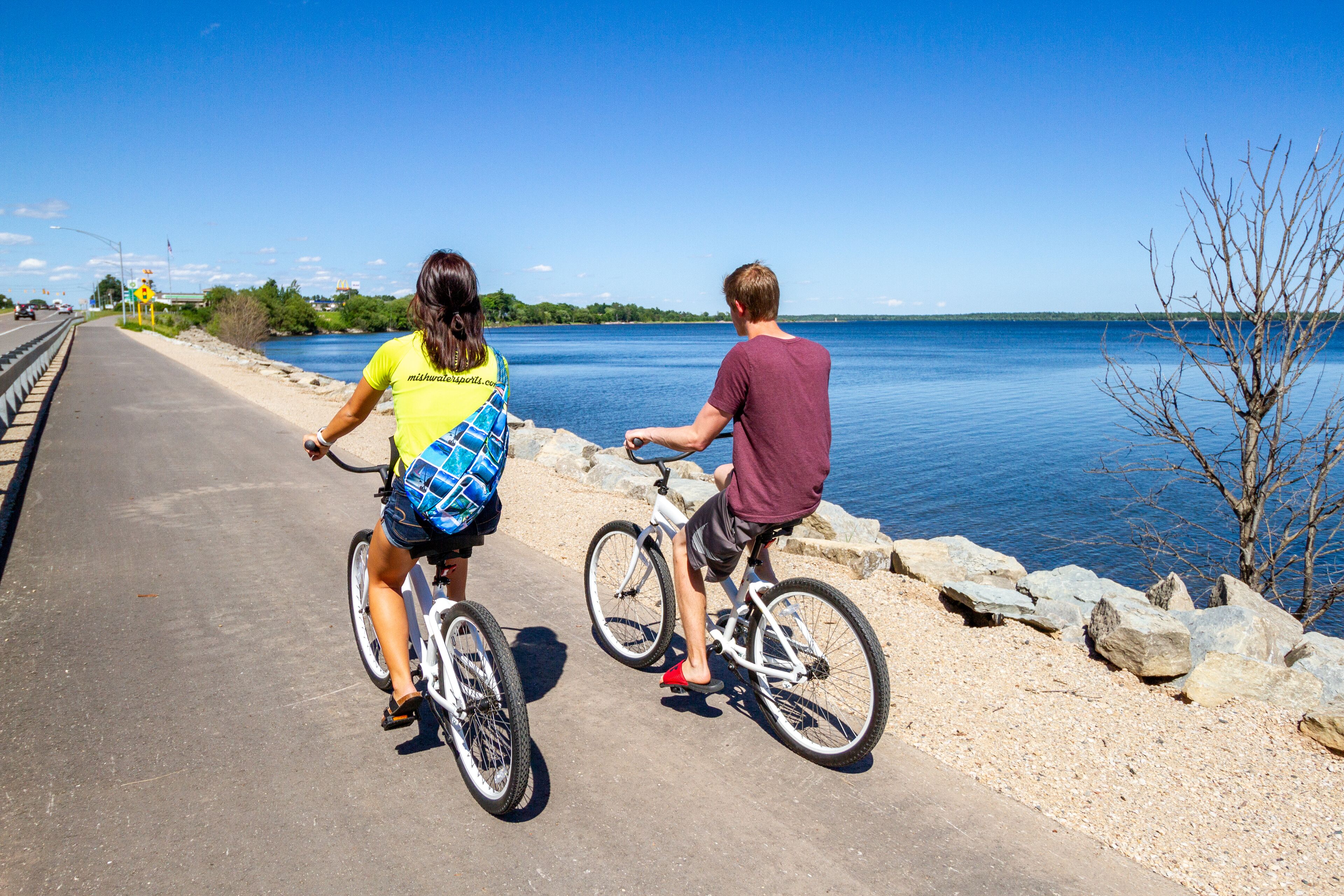 Two people riding bicycles on a paved path beside a body of water.