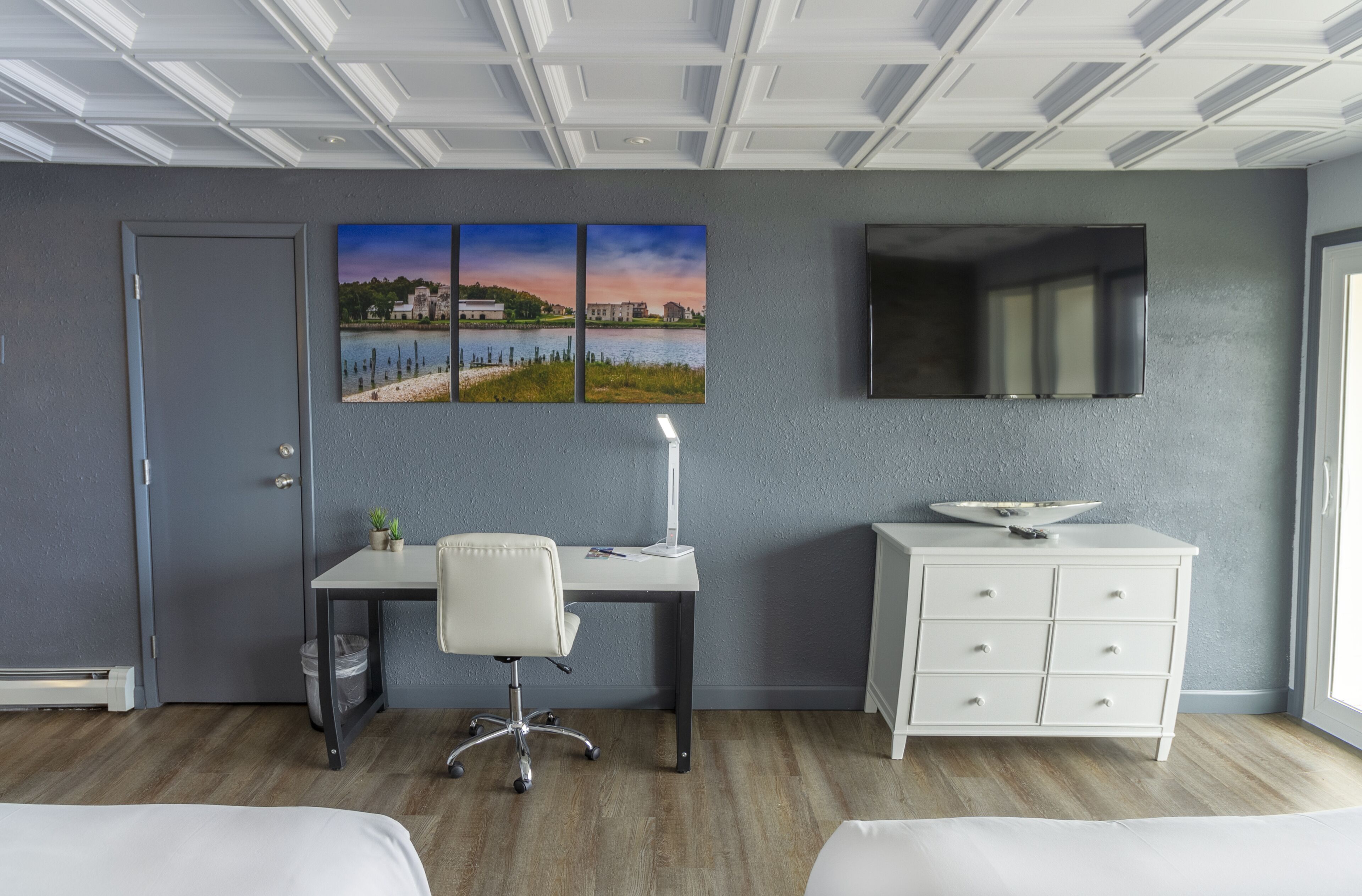 A hotel room with a bed, a desk with a chair, a dresser, a flat-screen TV, and a wall with three landscape paintings.