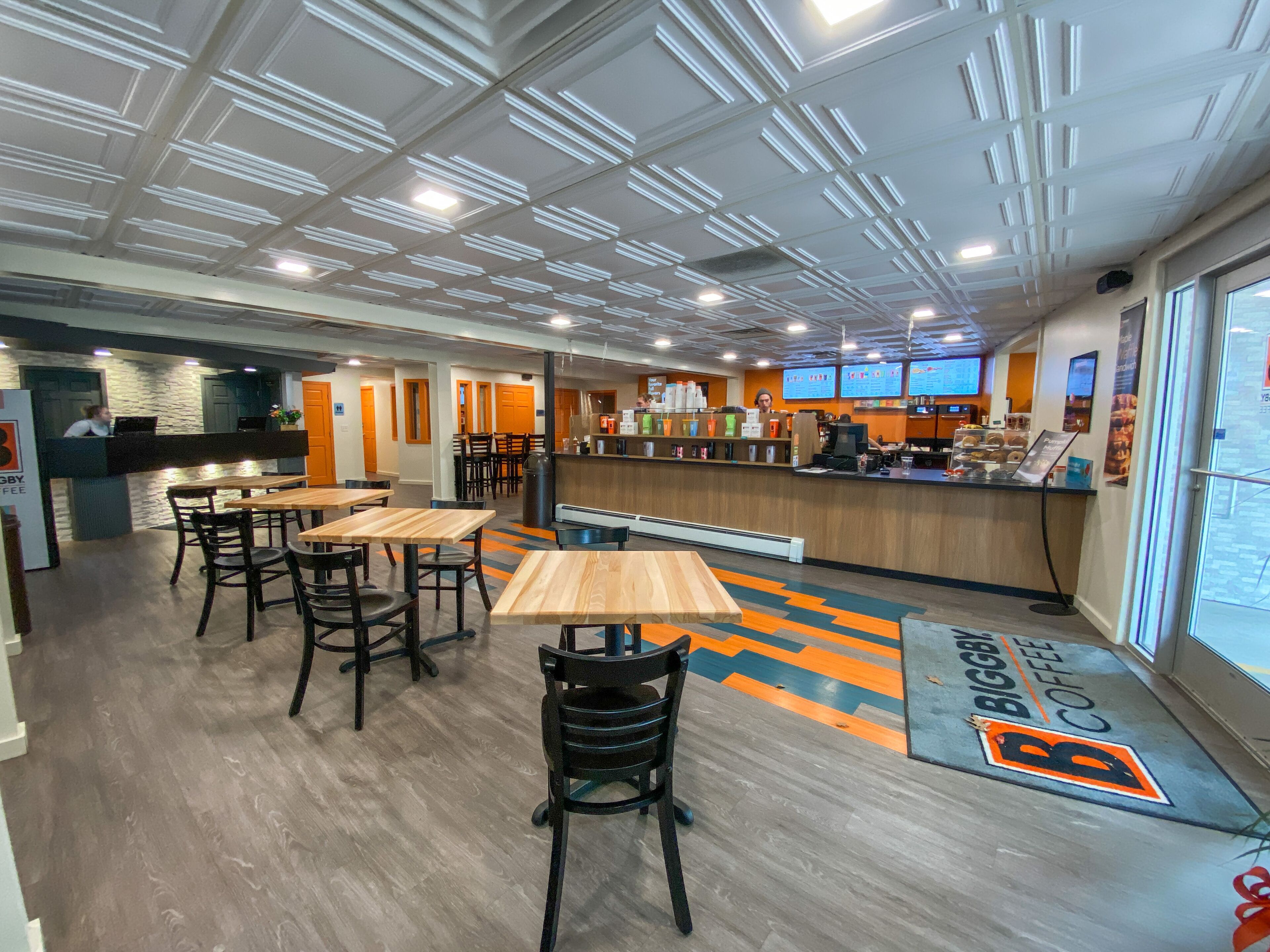 A modern coffee shop interior with wooden tables and chairs, a counter with a menu display, and a patterned floor.