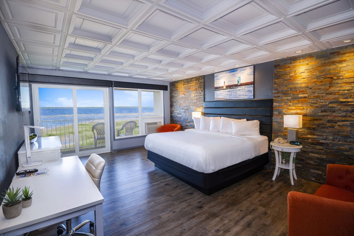 A modern hotel room with a large bed, a desk with a chair, a small table with a lamp, and a view of the sea through large windows.