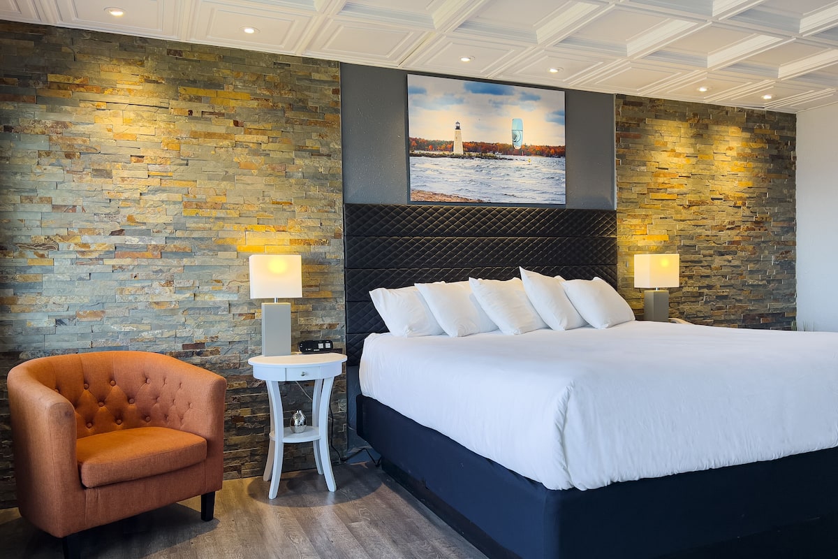 A modern hotel room with a large bed, a tufted armchair, a small side table, and a stone wall.