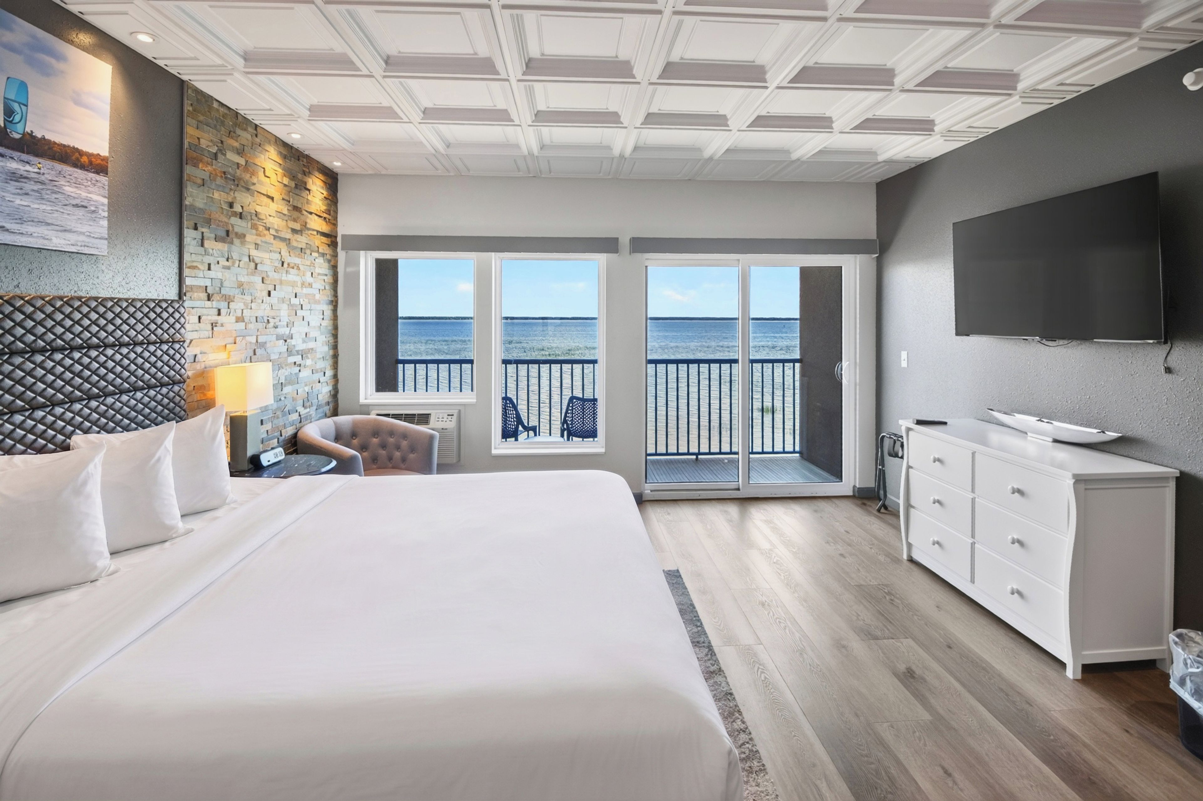 A hotel room with a large bed, a TV, a desk, and a view of the sea through the balcony doors.