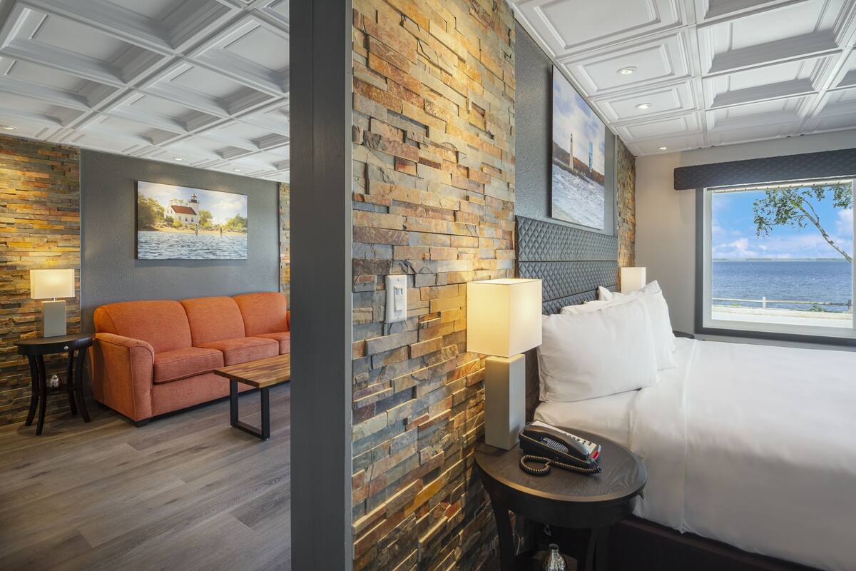 A hotel room with a stone wall, a sofa, a bed, a nightstand, and a view of the sea.