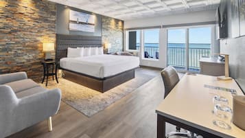 A hotel room with a large bed, a desk, a chair, a TV, and a balcony with a view of the sea.
