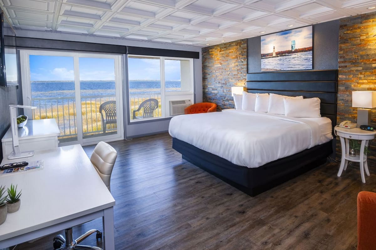 A modern hotel room with a large bed, a desk with a computer, a chair, and a view of the beach.