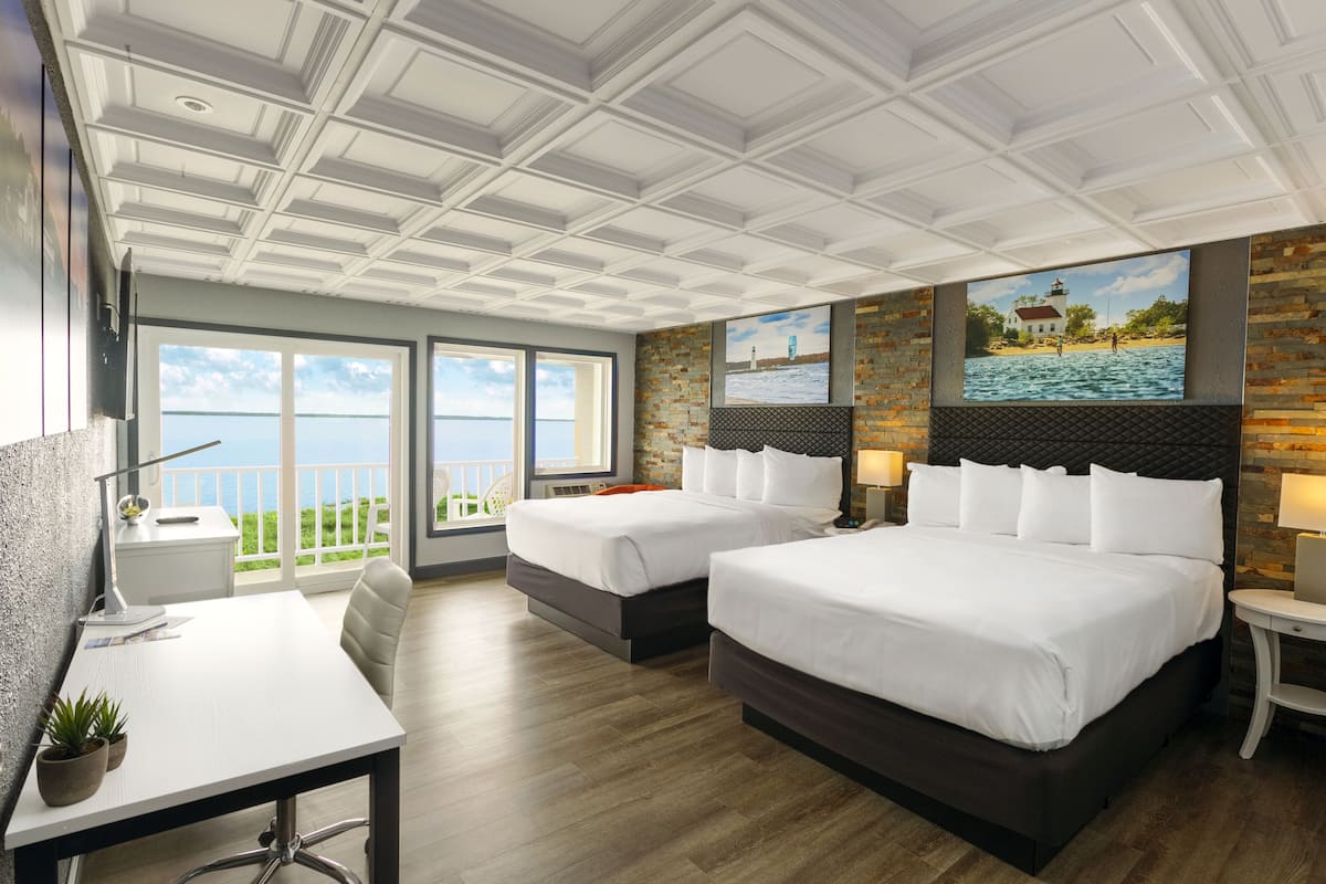 A hotel room with two beds, a desk, and a large window offering a view of the sea.