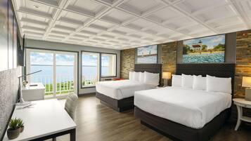 A hotel room with two beds, a desk, and a large window offering a view of the sea.