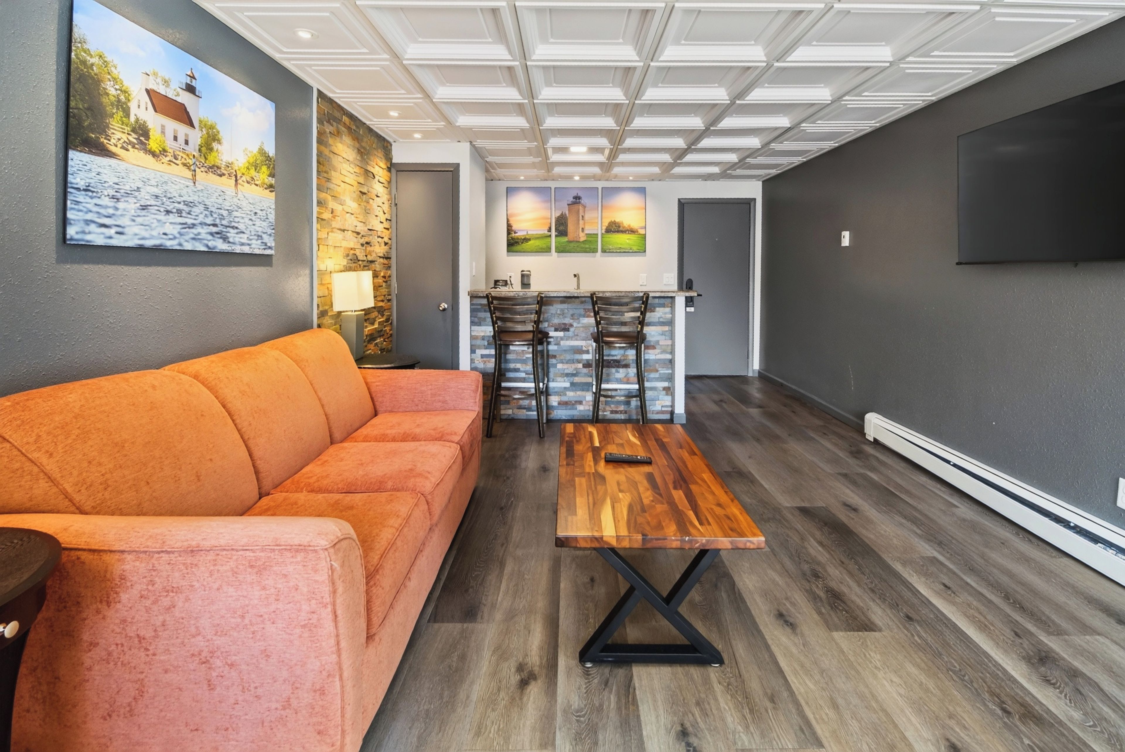 Un salon moderne avec un canapé, une table basse en bois et un coin cuisine doté d’un mur de crédence en pierre et de tabourets de bar.
