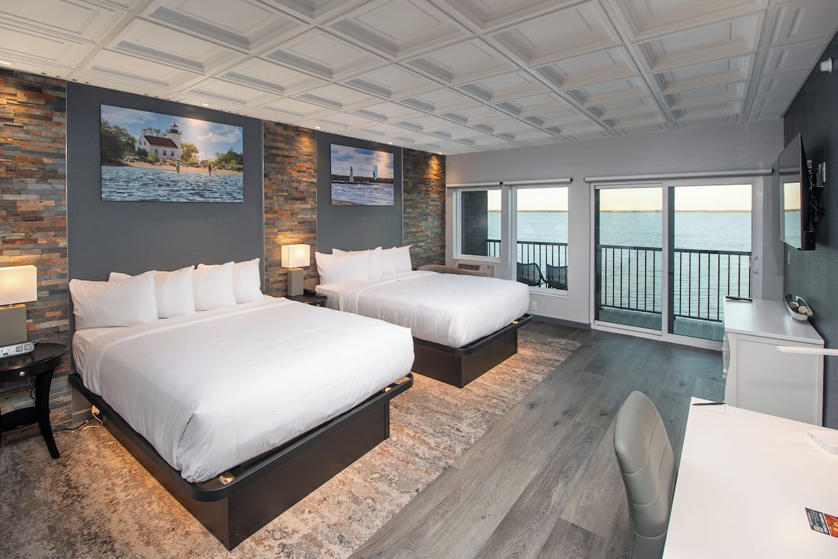 A modern hotel room with two beds, a desk, and a balcony with a view of the sea.