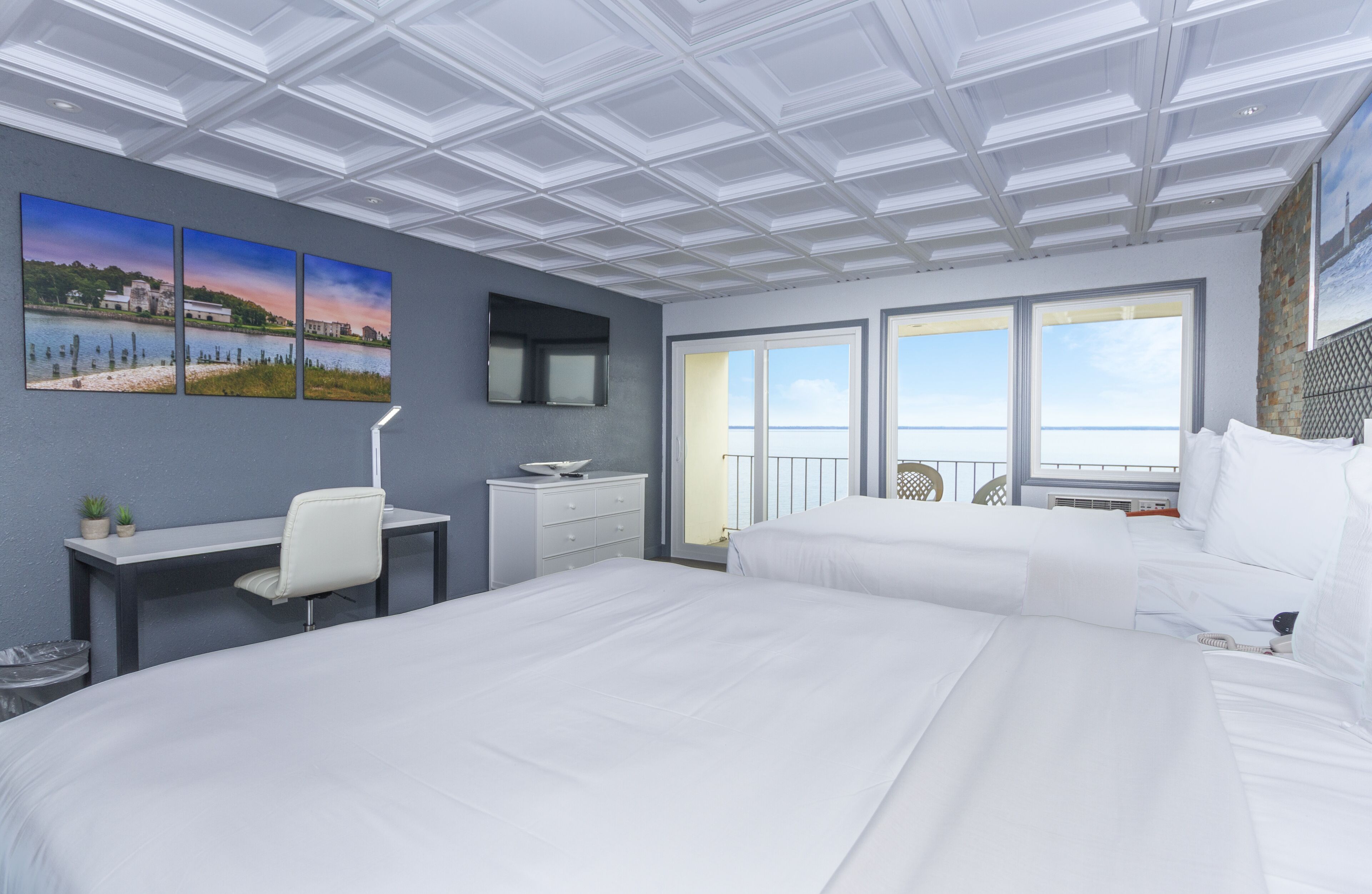 A hotel room with a large bed, a desk with a chair, a small table, and a TV. There are three paintings on the wall, a window with a view, and a balcony with a railing.