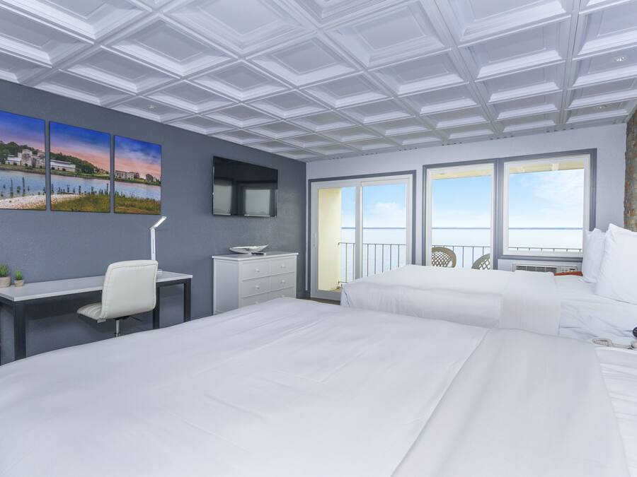 Habitación de hotel con una cama grande, un escritorio con una silla, una mesita pequeña y un televisor. Hay tres cuadros en la pared, una ventana con vistas y un balcón con barandilla.