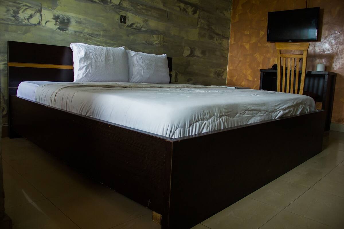 A large bed with a wooden headboard, a flat-screen TV mounted on the wall, and a nightstand with a lamp.