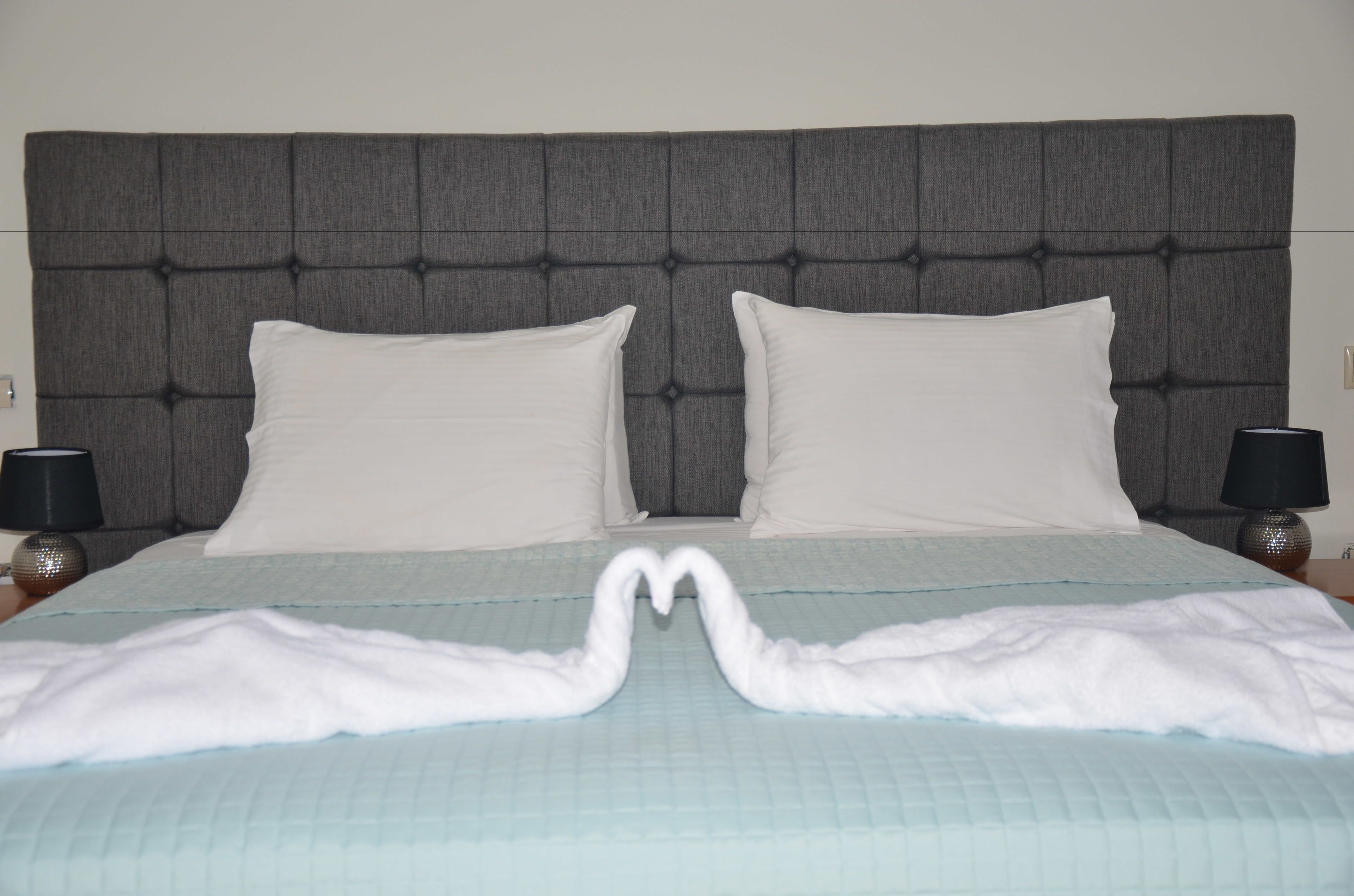 Ein Bett mit einem gepolsterten Kopfteil, zwei weiße Kissen und eine herzförmige Handtuchanordnung auf einer gesteppten Bettdecke.