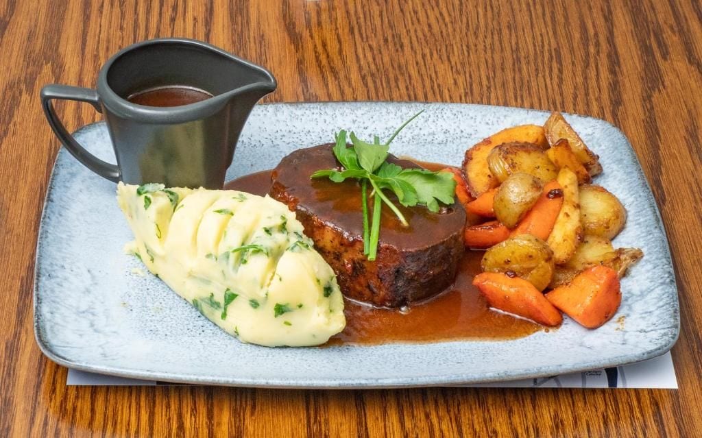 A plate of food with a meat patty, mashed potatoes, and roasted vegetables, accompanied by a gravy cup.