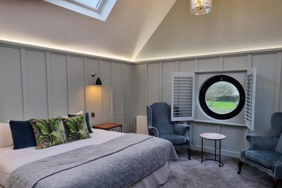Ein Schlafzimmer mit einem großen Bett, zwei Sesseln, einem kleinen Tisch, einem runden Fenster mit Fensterläden und einer Wandlampe.