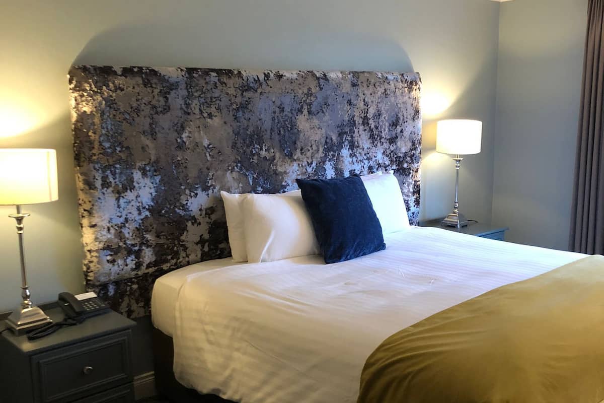 Ein Hotelzimmer mit einem großen Bett, zwei Nachttischlampen, einem Nachttisch mit Telefon und einem Kopfteil mit strukturierter Gestaltung.