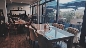 Un comedor moderno con una mesa de vidrio y sillas de cuero, ventanas amplias que ofrecen vistas al exterior, y una variedad de objetos decorativos en estanterías.