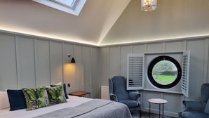 Un dormitorio con cama, dos sillones, una mesita, un ventanal con contraventanas y un tragaluz.