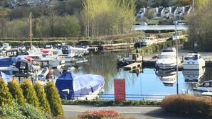 Una marina con barcos amarrados, un muelle pequeño y una colina con casas al fondo.