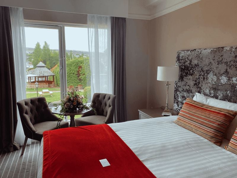 Ett hotellrum med en säng, två fåtöljer, ett litet bord med en vas och utsikt över en lusthus genom stora fönster.