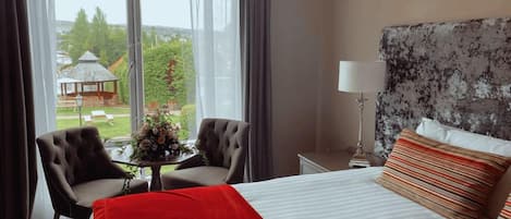 A hotel room with a bed, two armchairs, a small table with a vase, and a view of a gazebo through large windows.