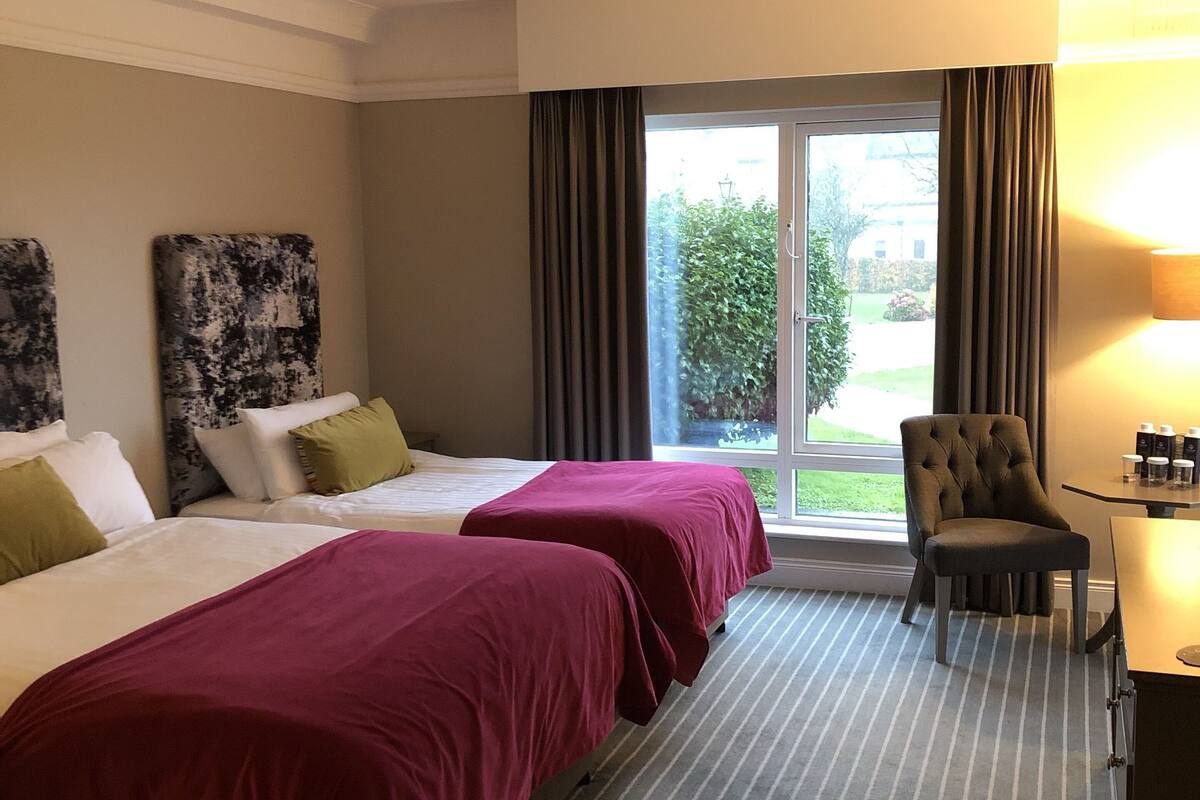 Ein Hotelzimmer mit zwei Betten, einem Sessel, einem Schreibtisch und einem Fenster mit Vorhängen.