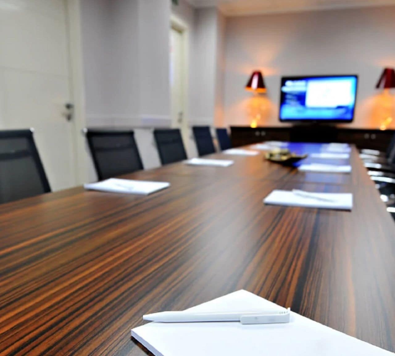 A conference room with a long wooden table, chairs, a laptop, and a television screen in the background.