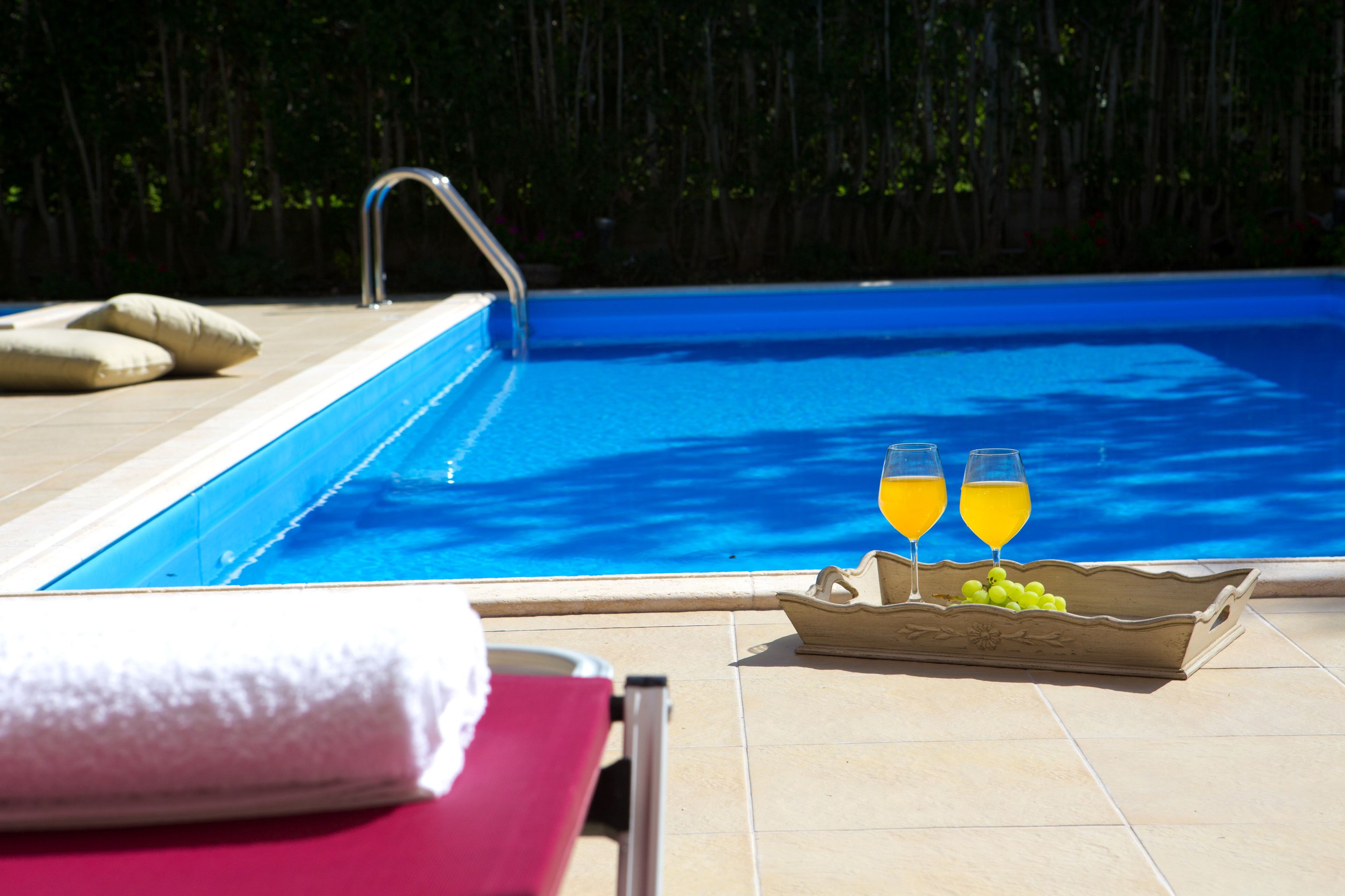 Une piscine avec une terrasse carrelée, un fauteuil de relaxation, une serviette et un plateau avec des verres de jus d’orange et des raisins.