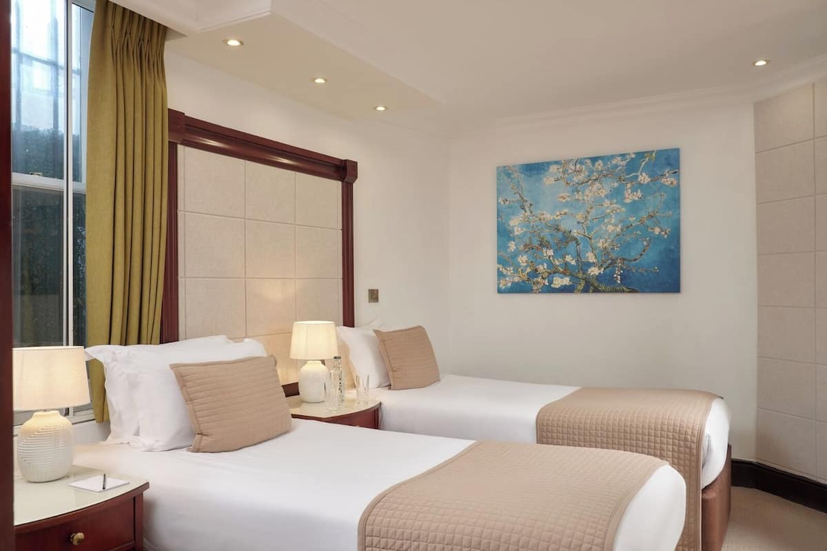 Una camera d'albergo con due letti, un'ampia finestra con tende, un quadro raffigurante un albero e un comodino con una lampada.