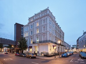 Un edificio a più piani con uno stile architettonico classico, caratterizzato da un ingresso ben visibile con un cartello, situato su una strada con auto parcheggiate.