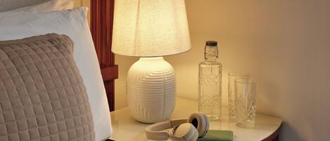 A neatly made bed with a quilted pillow, a bedside lamp, a bottle, glasses, and a pair of headphones on a bedside table.