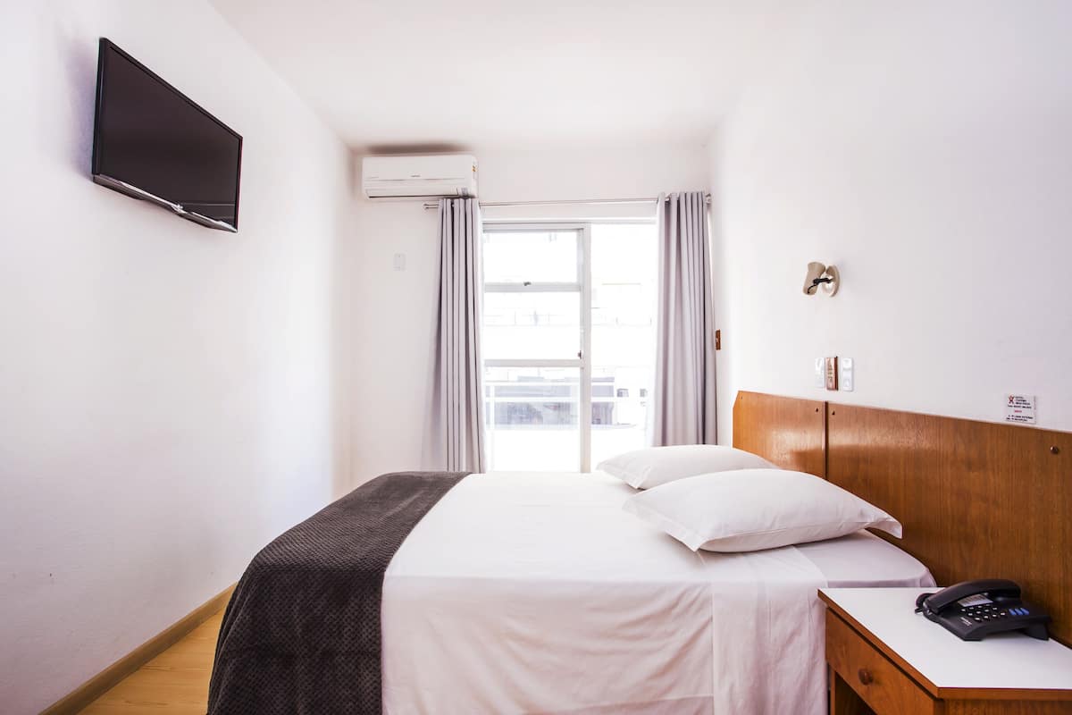 Ett hotellrum med en säng, ett nattduksbord med en telefon, en tv som är monterad på väggen och ett fönster med gardiner.