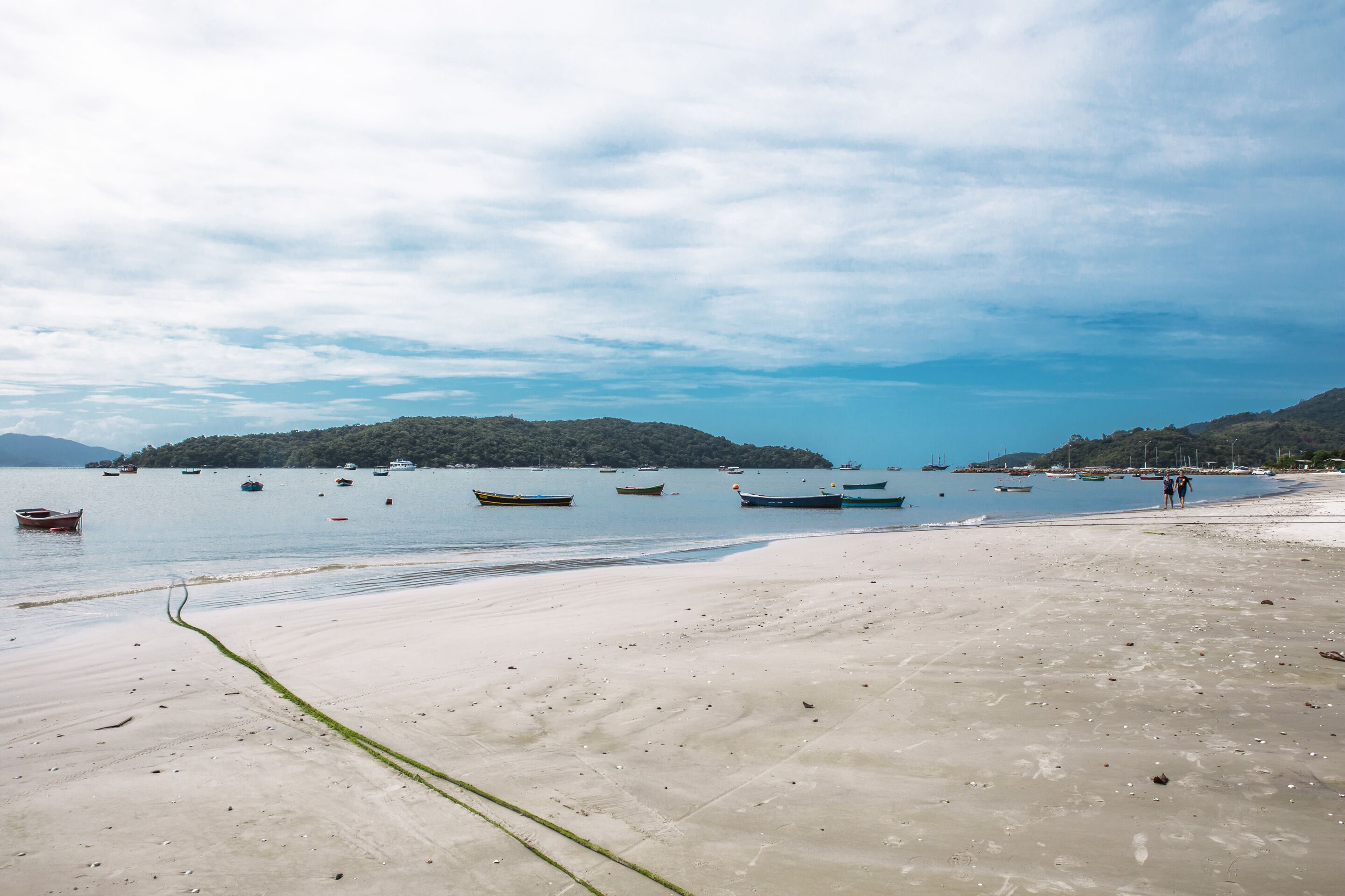 A beach with boats anchored in calm waters, a sandy shore, and distant hills.
