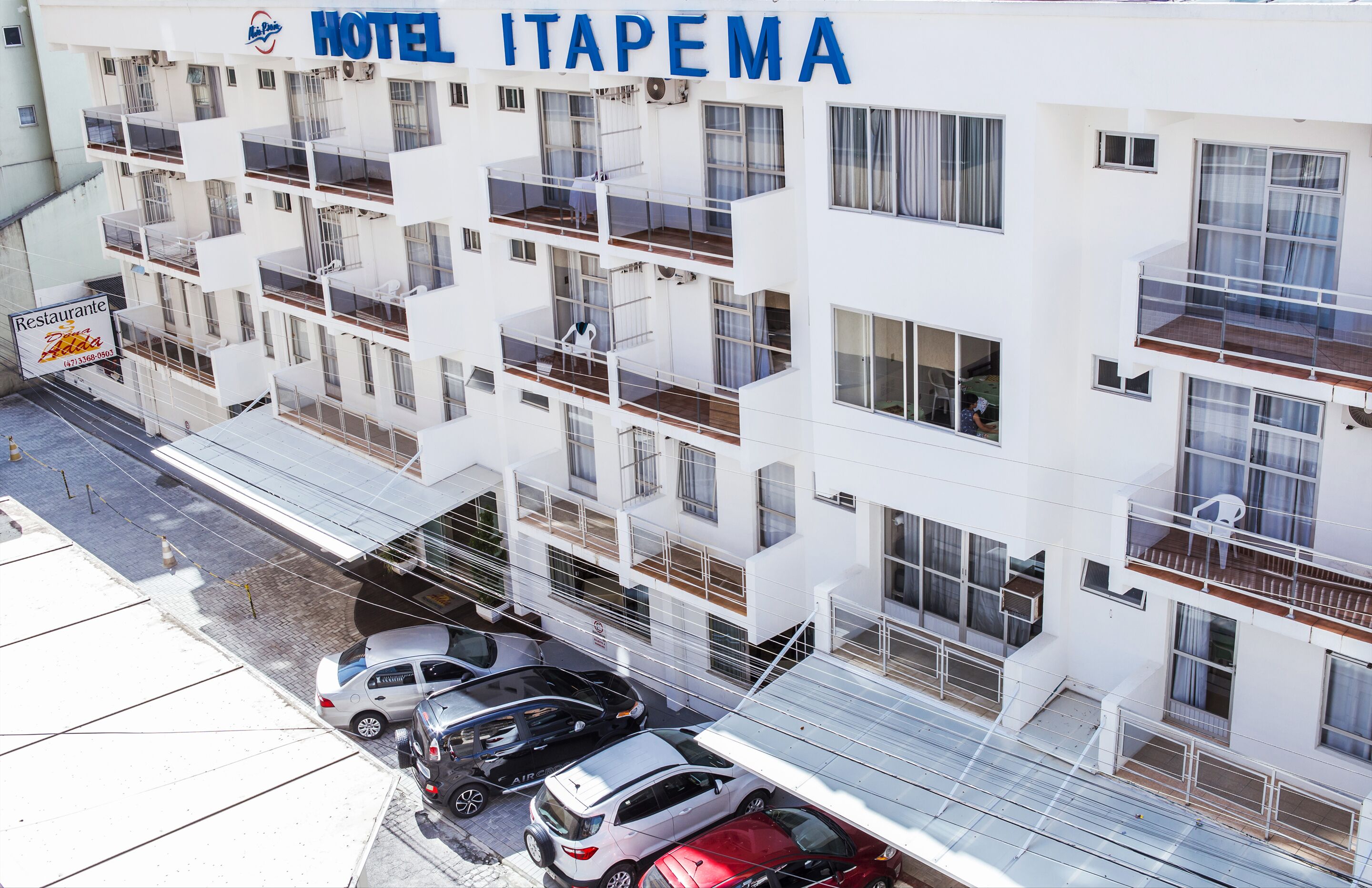 Un hotel de varios pisos con balcones, un letrero de restaurante y varios automóviles estacionados.