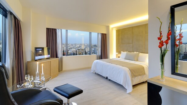 Una habitación de hotel moderna con una cama grande, un televisor en un mueble de madera, una mesita de cristal con copas de vino y vistas a la ciudad a través de amplios ventanales.