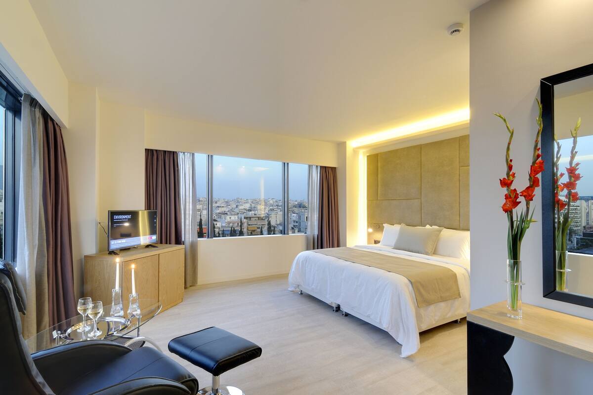 A modern hotel room with a large bed, a TV on a wooden cabinet, a glass table with wine glasses, and a city view through large windows.