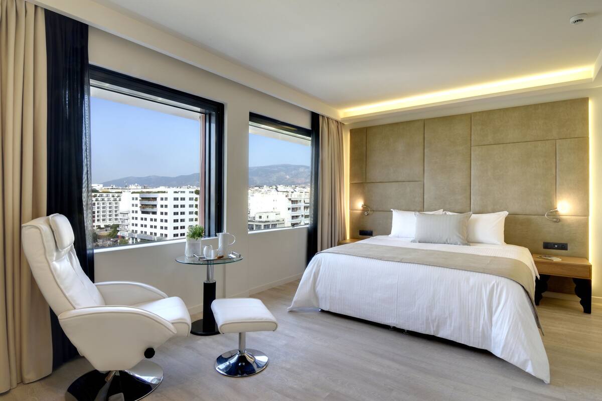 A modern hotel room with a large bed, a sitting area with a chair and a small table, and large windows offering a city view.